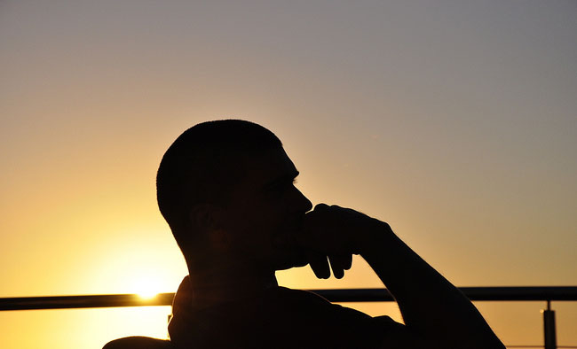 This is an image of a silhouette of a  person sitting in a pondering position with his hand rested on his chin, staring into the distance against a background of a setting sun. He appears to be deep in thought.