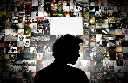 This is an image of a silhouette of a person pondering in front of a wall that displays a mosaic of pictures of people engaged in different activities at school, at work, at home, and in the community. In the centre of the collage is a blank rectangle. The person is thinking about what pictures of her or him might be there in the future.