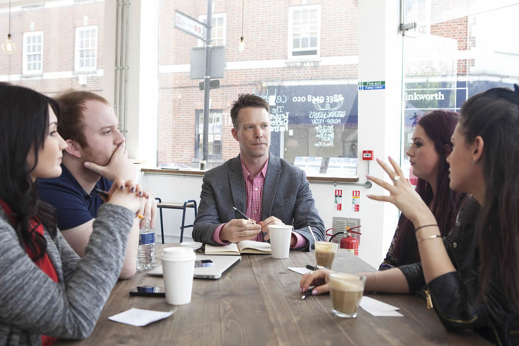 This is an image of five people both men and women, in casual business attire, sitting around a table in a coffee shop participating in a group meeting.
