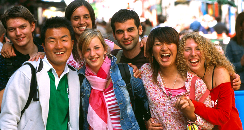 This is an image of a group of people with varied ethnicities, posing together for a group photo. Individuals are smiling, with their arms around each other's shoulders. They appear to be very happy.