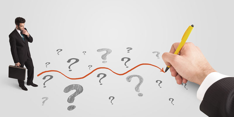 This is an aerial view of a young man in a business suit holding a briefcase and standing on the left side of a completely white surface. He is looking downward and has his free hand on his chin, appearing to be deep in contemplation. On the right side of the image is a large hand holding a pen and drawing a wavy red arrow from the man&rsquo;s feet  through a series of varying sized question marks that have been drawn in pencil.