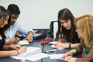 This is an image of four young adults seated around a table. Each has some form of notebook in front of them and is holding a writing utensil. They are all focusing intently on one of the group members who is sharing her knowledge with them. The three are engaged, focused, and taking notes.
