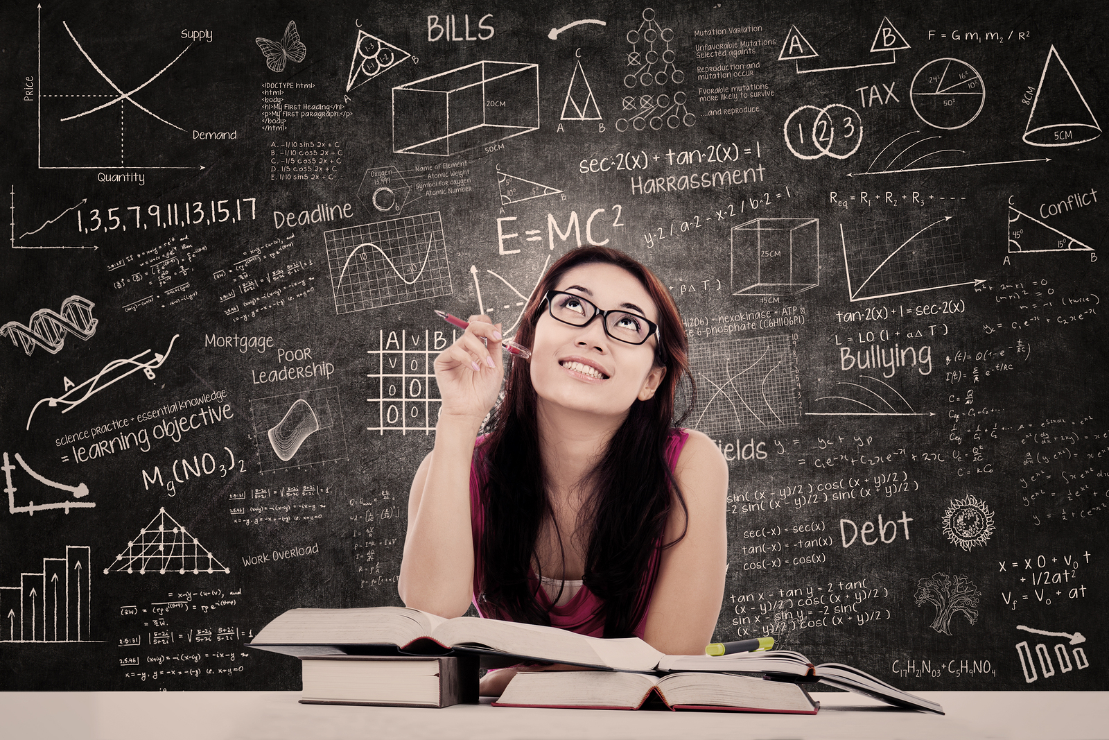 This is an image of young adult woman seated at a table that contains a number of books, opened and closed. Behind her is a chalkboard that is covered with diagrams, charts, graphs, mathematical formulas, and concepts. She is holding a pen, smiling and looking upward. She is focused and engaged in reflection about all she has learned.