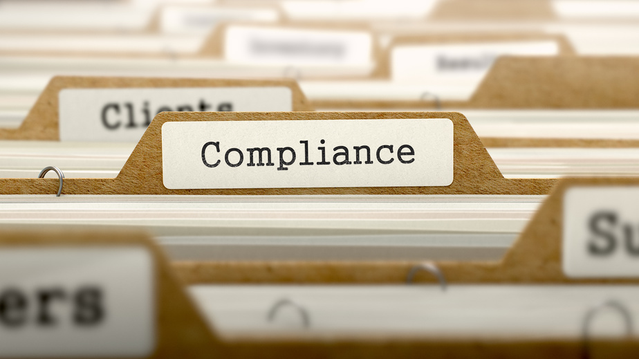 This is an image of the tops of a number of labelled files in a file cabinet with one pulled slightly upward. This file contains the label, ‘Compliance.’