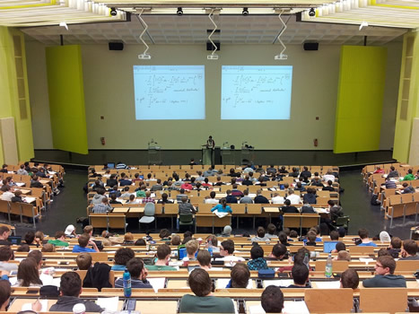 This is an image of a large auditorium taken from the back. Most of the seats are filled with students who are listening to math lecturer who stands behind a lectern. Two projectors are casting large images of mathematical equations behind the lecturer.