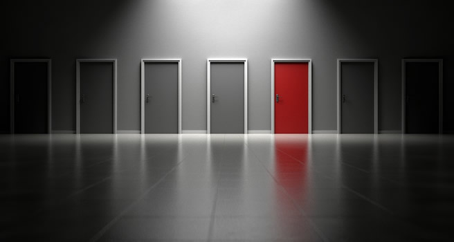 This is an image of an empty grey lobby with several doors that the end. The doors in the centre of have a spotlight on them, making them appear more prominent in the image. One of these doors is red, while all others in the image are grey.