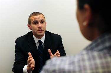 This is an image taken from behind an interviewer, showing an interviewee dressed in business attire. The interviewer looks on while the interviewee attempts to speak convincingly through pronounced facial expression and hand gestures. He does not have a portfolio or any artifacts to provide evidence his claims.
