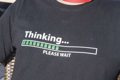 This is an image of the front of a person&rsquo;s dark grey T-shirt. A half-filled green status bar (like one would see on a computer) is printed on the shirt as well as white lettering that reads &lsquo;Thinking dot dot dot, please wait.&rsquo;