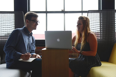 This is an image of two young adults seated on cushioned seats on opposite sides of a very small table. One is holding a pad of paper and a pen in his lap, while the other has her hands on a laptop that sits atop the desk between the two. Smiling, she demonstrates her online portfolio to the gentleman who looks onward with an intrigued look.