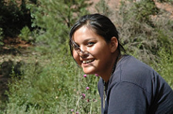 This photograph shows a young indigenous woman smiling, in a nature setting.