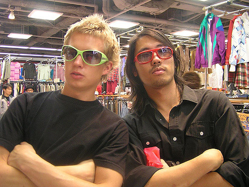 This photograph shows two young men in a store standing side by side, with arms crossed. They are both wearing sunglasses.