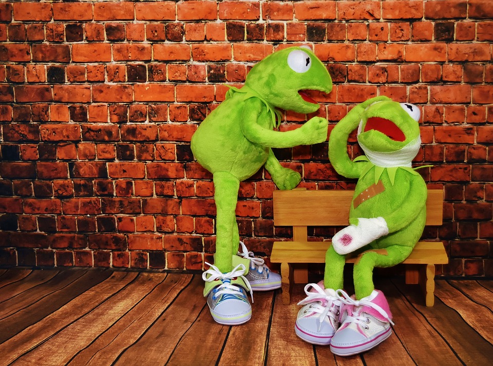 This is an image of two puppet Kermit the Frogs.  One is sitting on a bench bleeding with injuries while the other frog leans over it.
