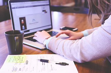 This is a photograph of a laptop, coffee mug, paper, pen and cell phone on a desk.
