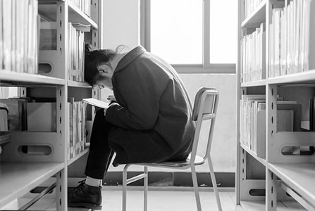 This is a black and white photograph of a girl sitting on a chair taking notes in a library.