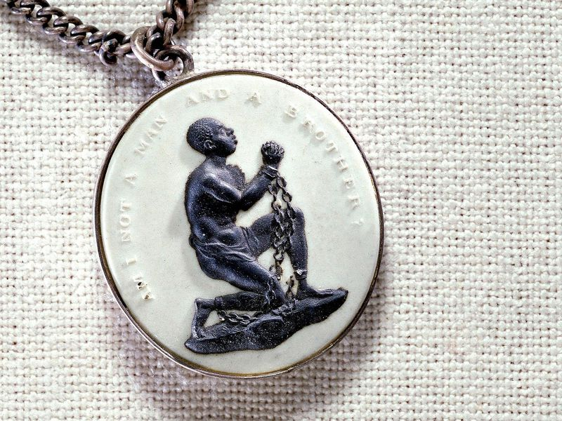 This is an image of a pendant, which appears on a necklace chain, shows a slave who is shackled and kneeling down. The words “Am I not a man and a brother” are written above the kneeling slave.