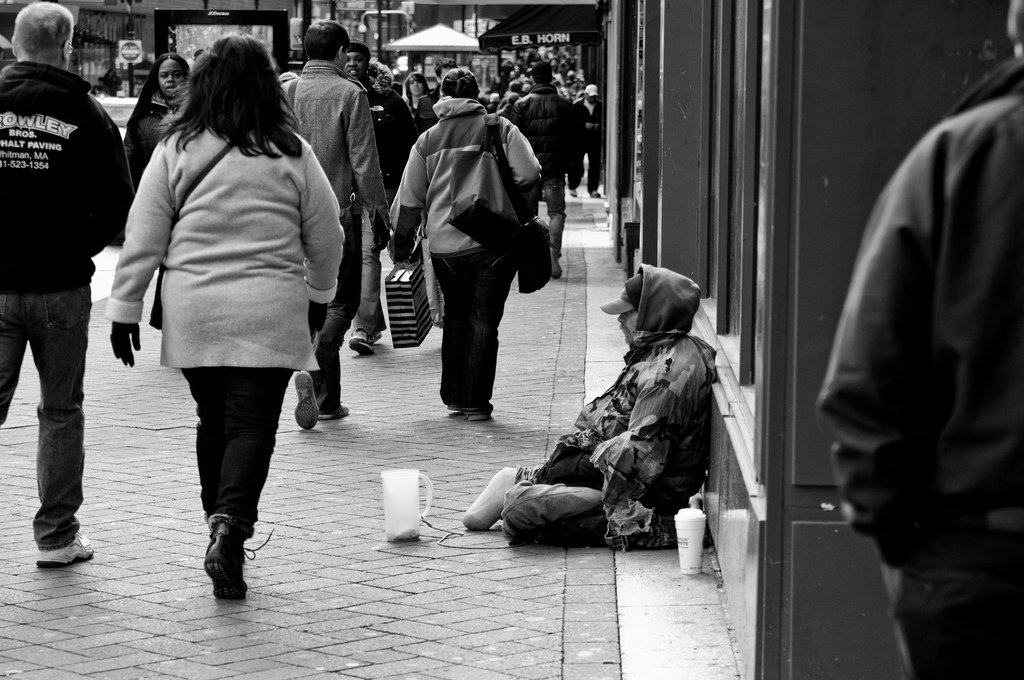 This is a picture of a homeless man sitting on the sidewalk with a container in front of him for people to donate money.  Lots of people are walking by him without even turning to look at him.