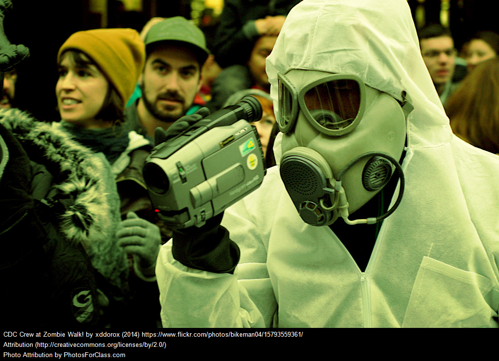 This is an image of a person in a crowd of people who is wearing a white protective suit and holds a video camera.