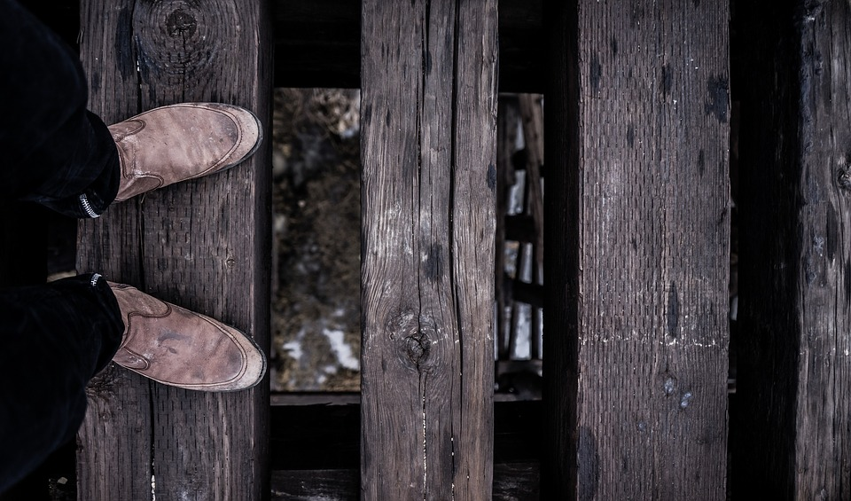 This is a close-up image of a pair of boots standing on the wooden slats of a bridge. Through the slats you can see the water, far below.