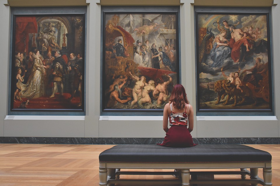 A young woman sits in a gallery, observing three large paintings.