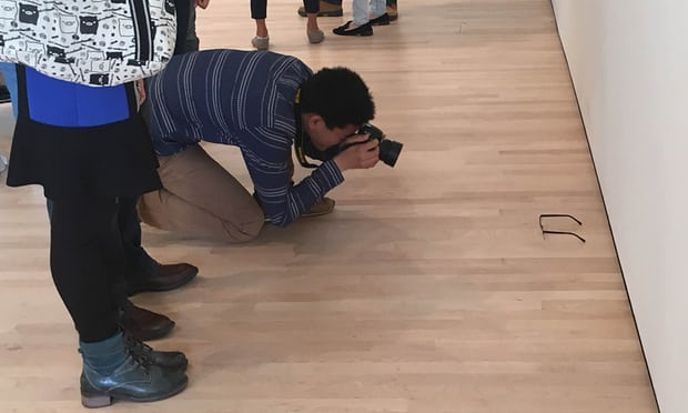 This is an image of a man taking a photograph of a pair of glasses on the floor of an art gallery.