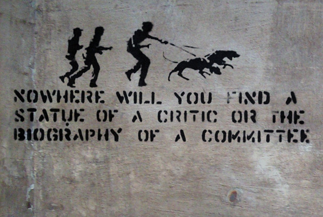 This is an image of an early artwork by Banksy. It features the silhouettes of men following bloodhounds in pursuit. Underneath the image is text that reads, “Nowhere will you find a statue of a critic or the biography of a committee.”
