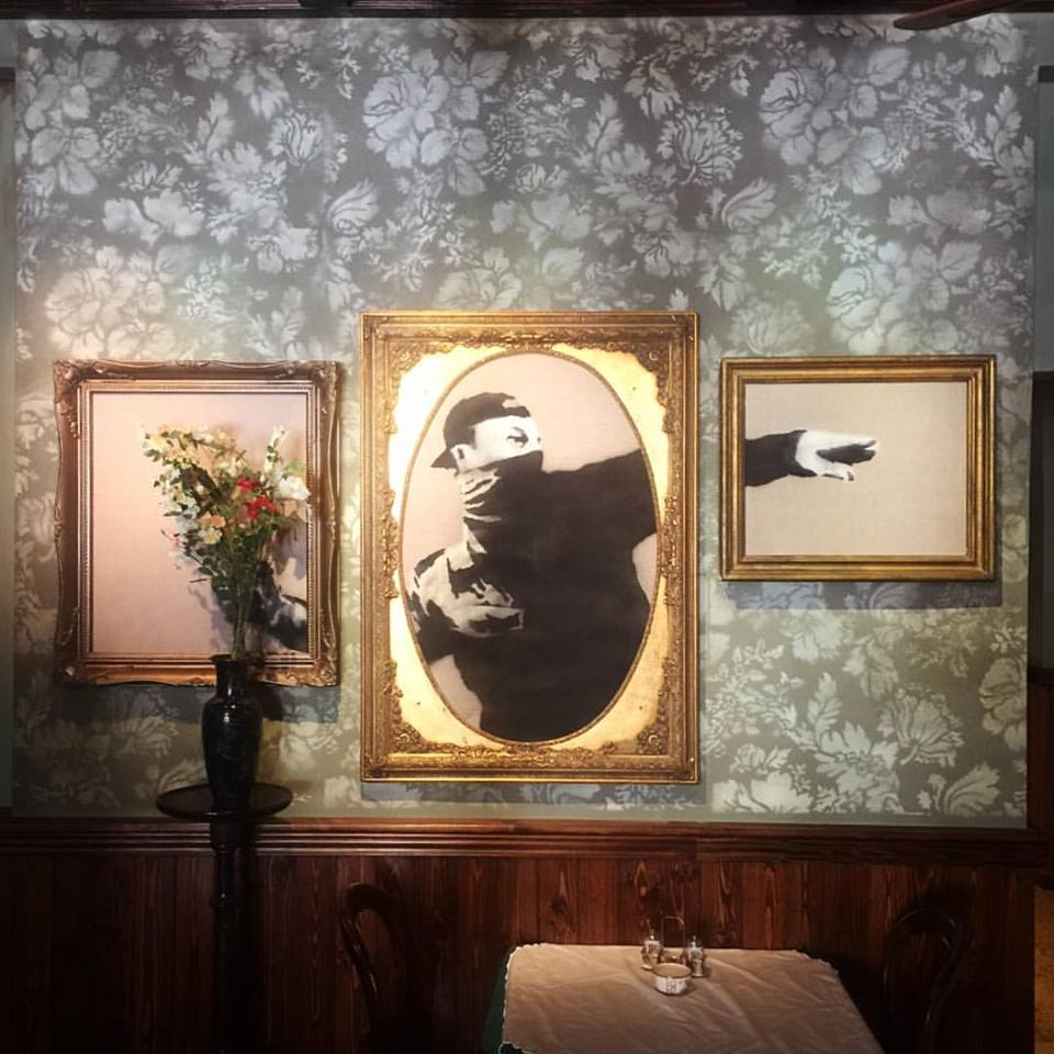 This is an image of one of Banksy’s famous murals of a protester throwing a bouquet of flowers, into three gentrified, gilt-framed paintings.