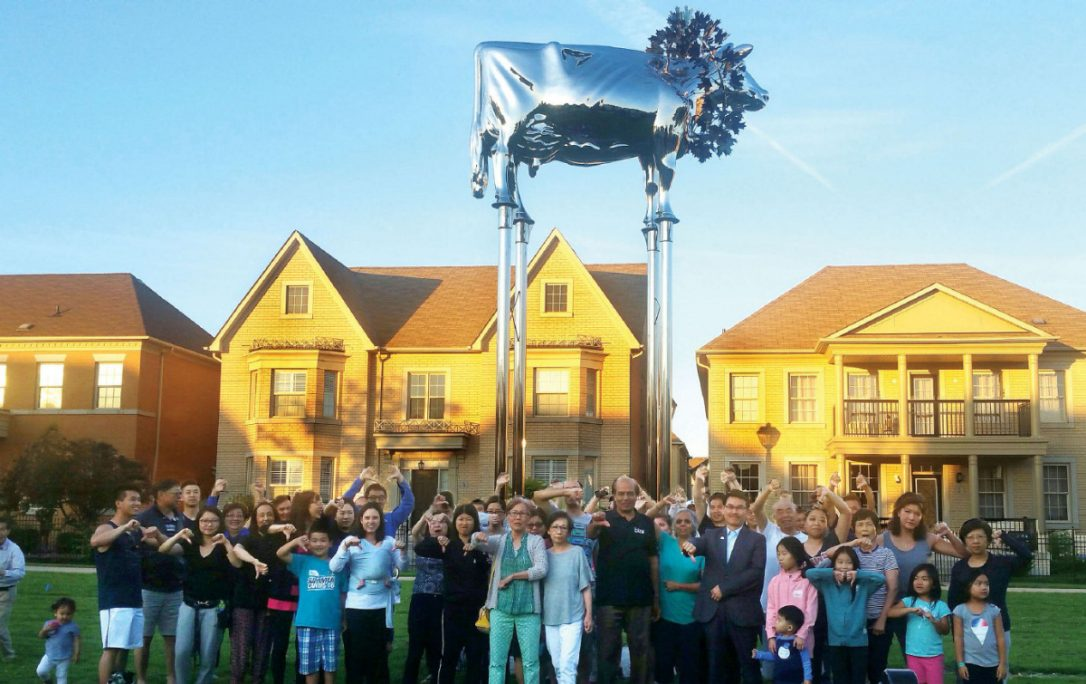 This is an image of a crowd of people, giving the thumbs down gesture to the statue of a large, silver cow, that is erected on stilts behind them.