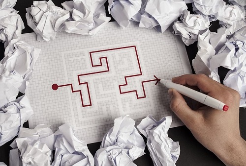 An image of a person completing a maze. There are several scrunched up pieces of paper around, indicating that they have tried several solutions.