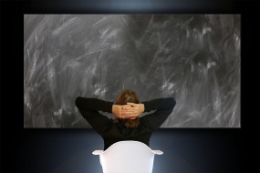 An image of a woman leaning back on her chair staring at a blank chalkboard, contemplating a solution to a problem.