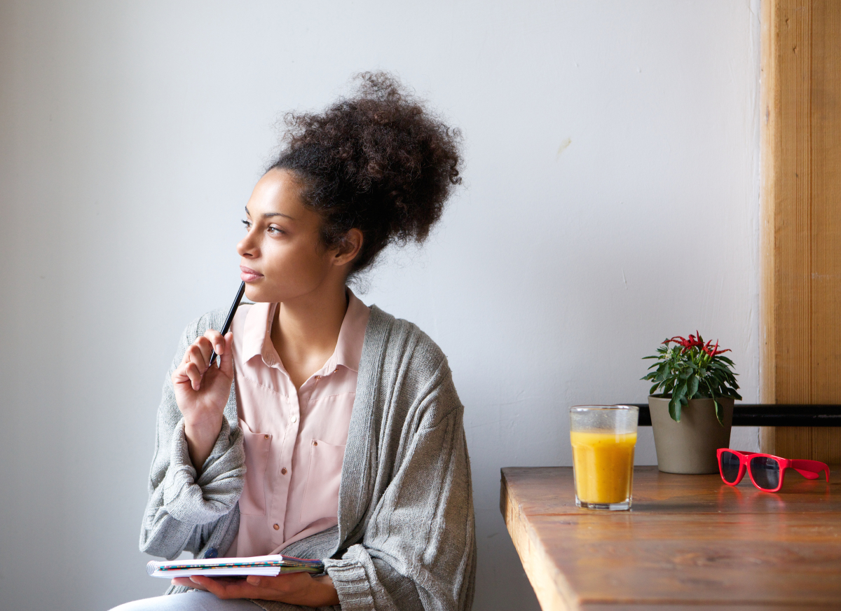 This is an image of a woman sitting at home with pen and paper thinking while working at a counter with a drink of orange juice.