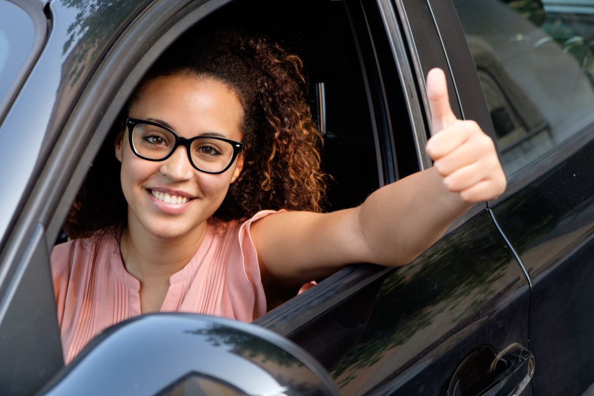 This is an image of a girl in the driver's seat of a car with thumbs up sticking out the window.