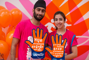 This is a picture of a young man and woman from a university in Thailand. They are standing in front of a wall with two hands crossing. They are wearing red t-shirts as part of a community run. They are both holding up giant hands. One hand has words written in Thai and the other hand has words written in English. The English words read, Strong hands stop violence against women and girls.