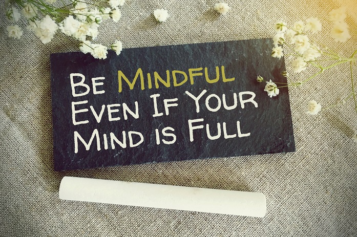 This is a picture of a chalkboard lying on a chair. There is writing on the chalkboard which says Be mindful even if your mind is full.  There are some white flowers at the top, and a piece of chalk is lying in front of the board.