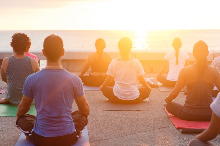   In this image, a yoga class is being held outdoors with nine people who are all facing a sunset as they sit on their mats in yoga poses.