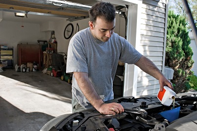 This is an image of a man who is standing outside his garage in front of his car and adding oil to it.