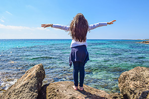 An image of a girl standing on a rock with her arms outstretched, looking at the ocean.