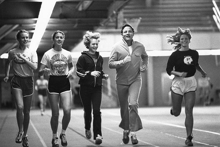 Man in track suit running with 4 female athletes towards camera on an indoor track.