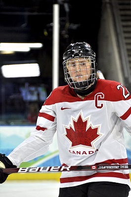 This female hockey player is wearing a red and white hockey jersey with a red Maple leaf in the centre  and the letter C on the left side of the shirt and and helmet with a full cage. She carries her hockey stick across her left hip. 