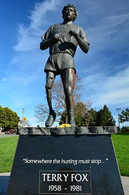This statue shows Terry Fox with his  prosthetic leg, dressed in athletic shorts, T-shirt and running shoes standing tall against the background of a blue sky. The caption on the base reads: Somewhere the hurting must stop...