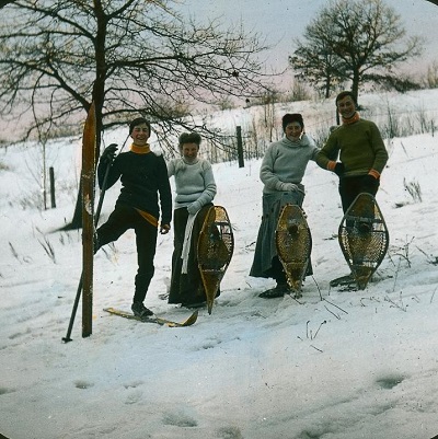 This early Canadian photograph shows two men and two women standing in a field covered with snow. They are all dressed in warm outdoor clothing, the men in pants and the women in long wool skirts and all wearing wool sweaters. The two women and one man are holding snowshoes, the other man is wearing skis.