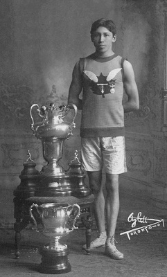 This old black and white photograph shows an athletic young man dressed in long athletic shorts, a sleeveless shirt with an emblem that bears the letter T, a maple leaf and wings on it, and is wearing vintage athletic shoes is standing beside two large trophies.