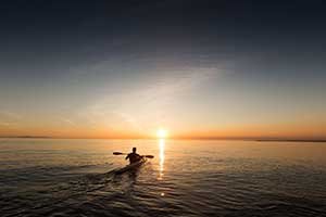 An image of a man paddling a kayak in the sunset.