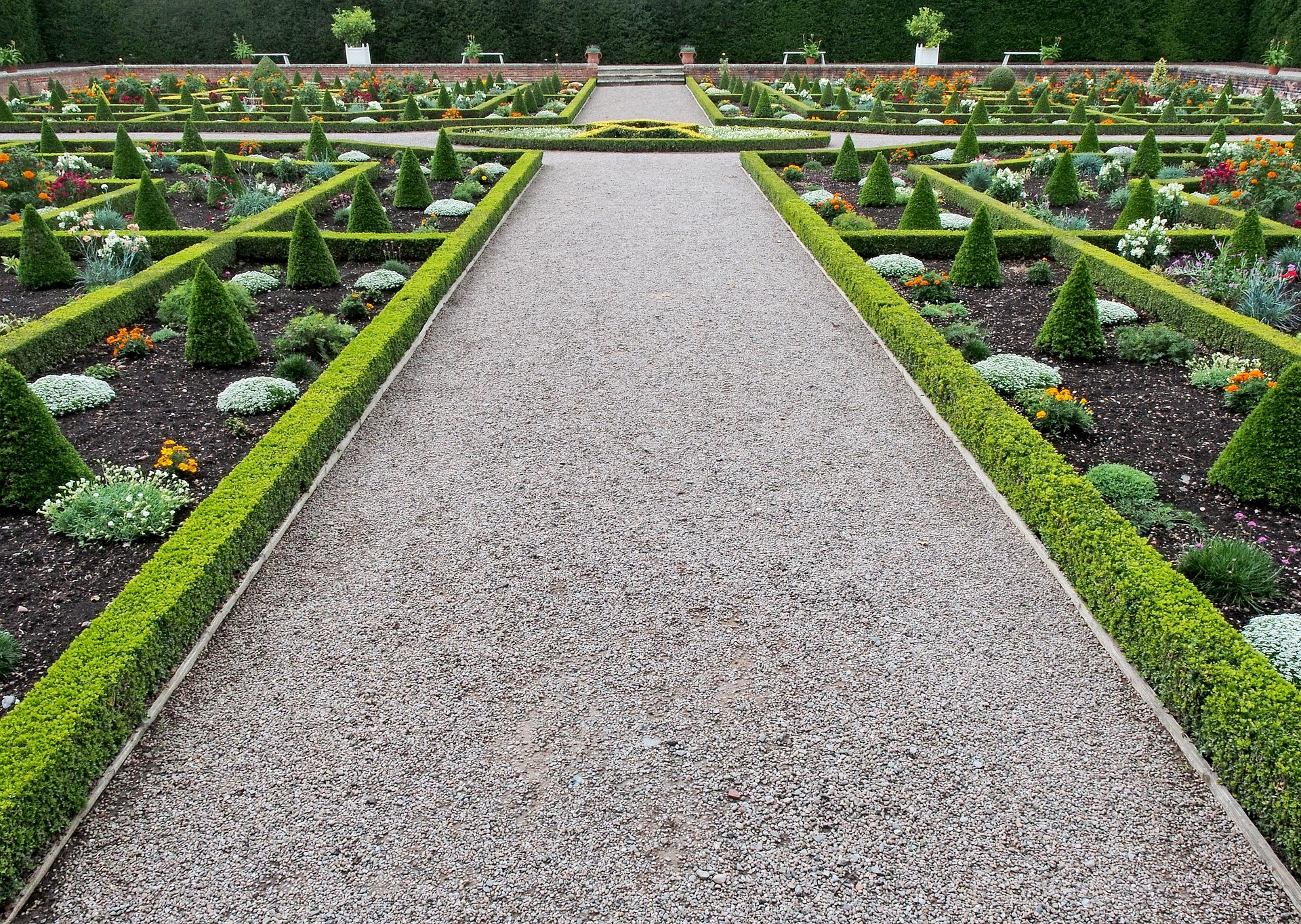 This is an image of a very manicured garden. A grey pebble walkway runs up through the centre of the image, with the gardens to the right and left of the path. The gardens have a border of squared off hedges and the gardens contain very evenly spaced plants flowers and shrubs.