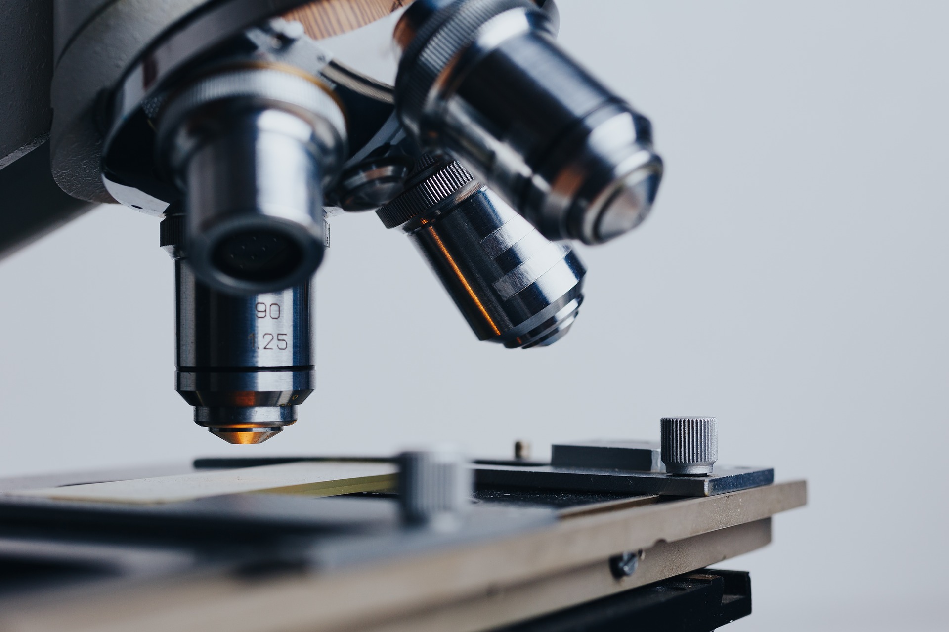 This is a closeup image of the nose piece of a compound light microscope. At the bottom of the image is the microscope stage, blurred in the foreground by in focus in the background. On the stage you can just see a glass microscope slide. The the top left of the image, there are four silver, cylindrical objective lenses attached to a round, silver convex dome. The lenses are arranged like the legs of a chair. The two lenses in the foreground are blurred, and the two lenses in the background are in focus. The background is grey.