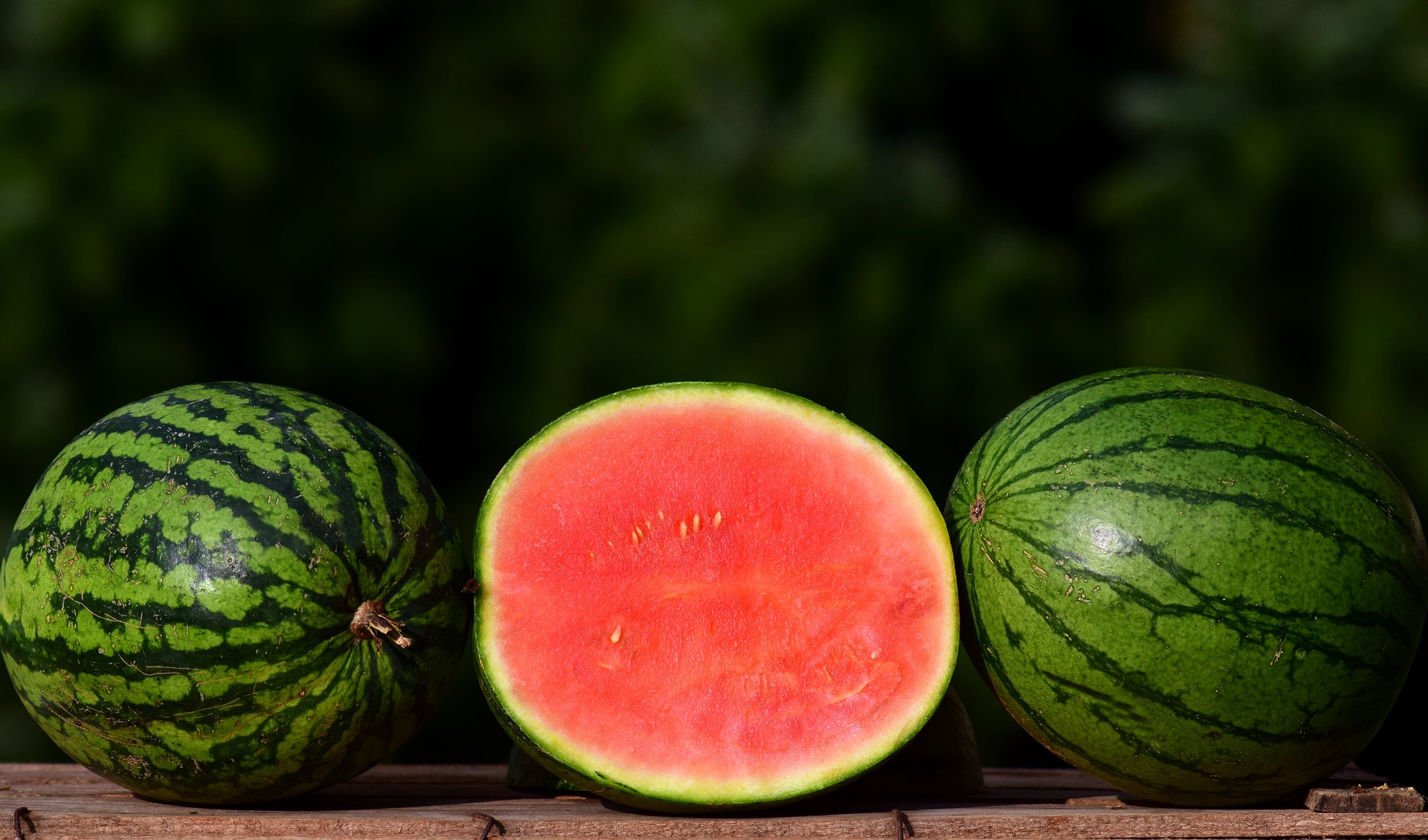 This is an image of three watermelons on a wooden table arranged in a row. The melons on the left and right of the image are whole, laying on their sides, and are green with darker green stripes running along them from top to bottom. The melon in the centre has been cut in half, with the cut, pink surface facing the viewer. There are blurred leaves in the background.