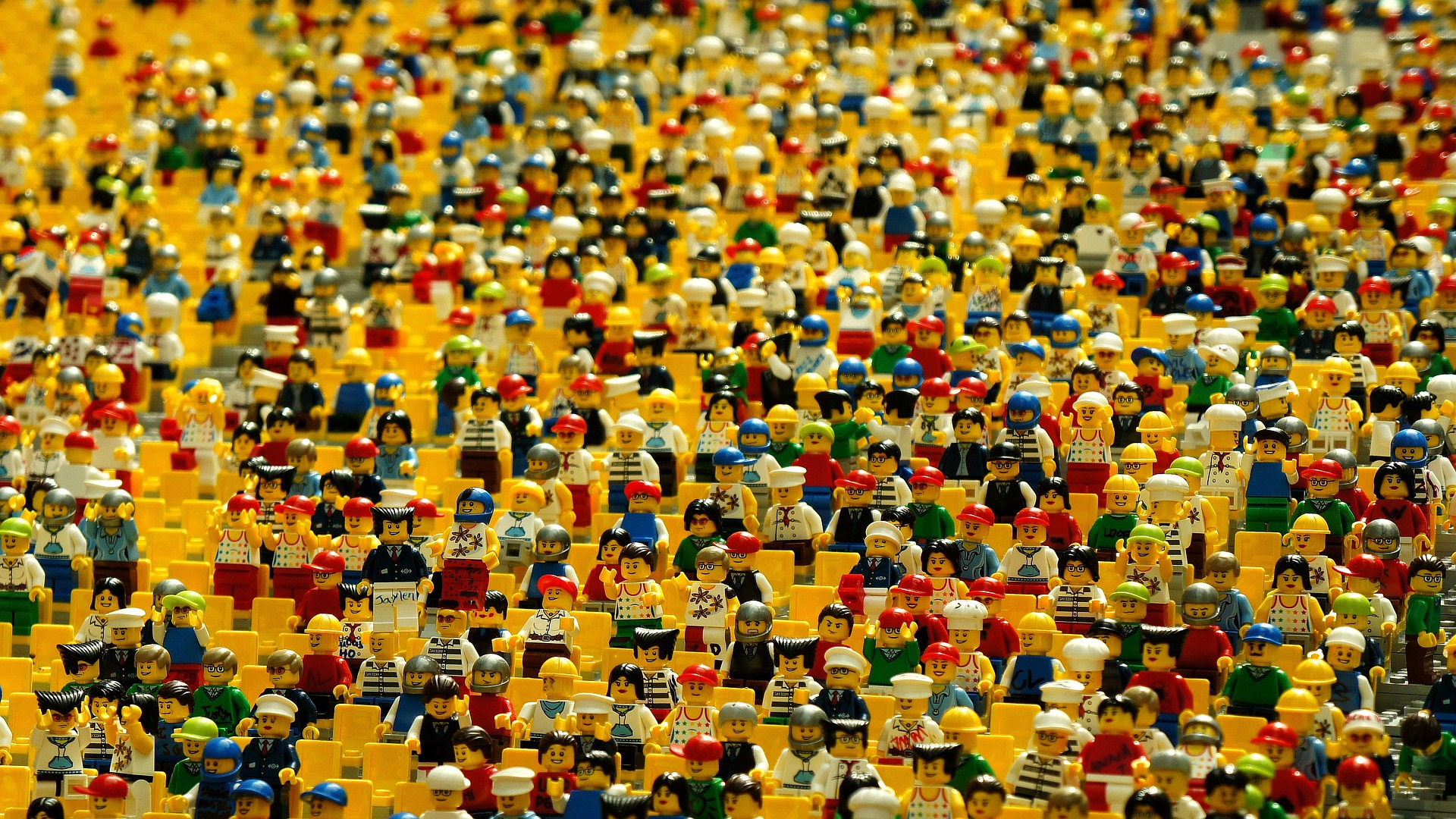 This is an image of a crowd of hundreds of Lego people. The crowd fills the entire image. A number of empty, yellow seats are scattered throughout the crowd. Many of the figurines are wearing hats.