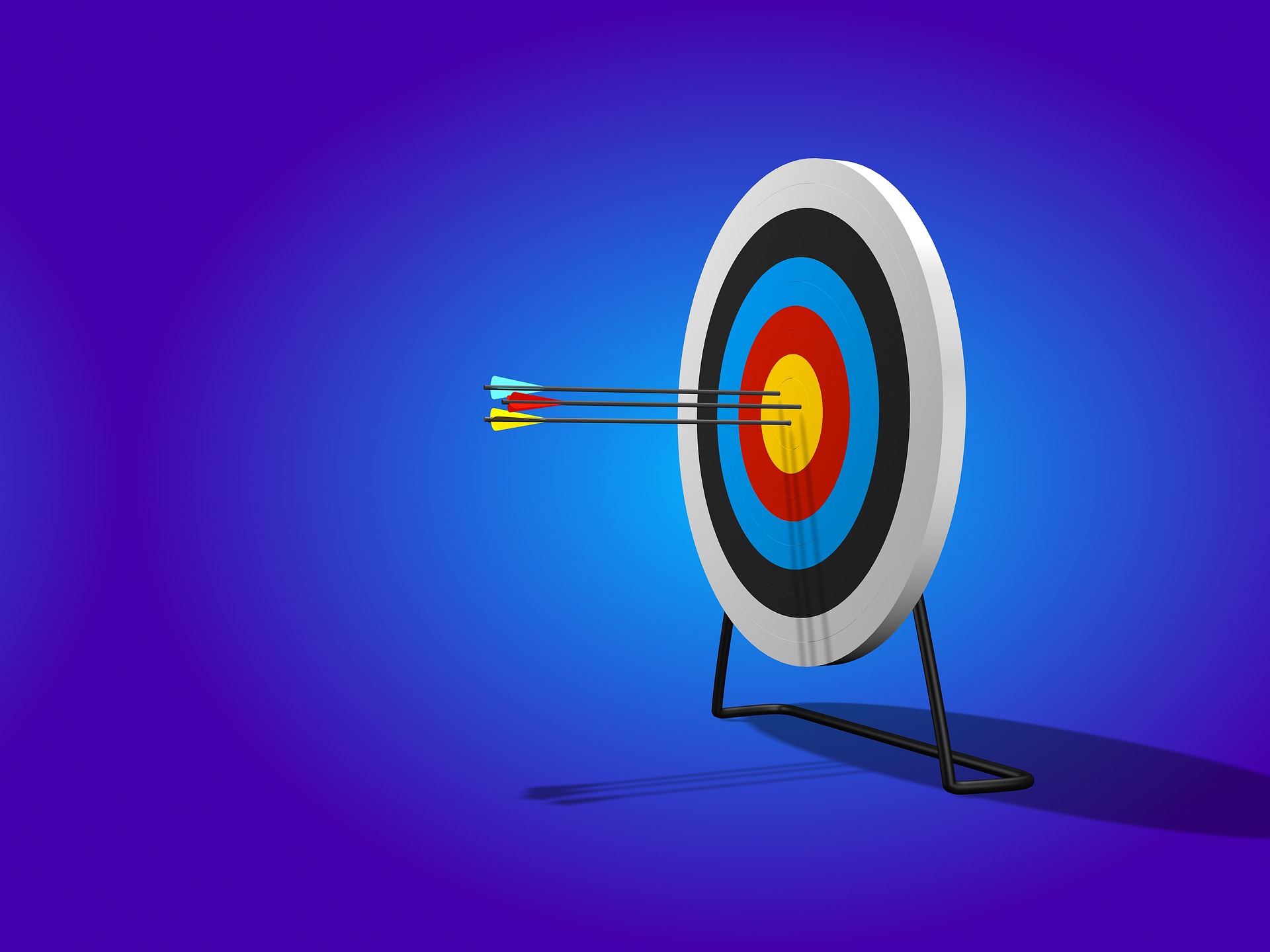 This is an image of an archery target on a stand against a blue background. The target is to the right of the image and is rotated to the left. Three arrows are in the centre of the target. One has yellow flights, another blue fights, and the last one red flights. There is a shadow of the target and the arrows below it.