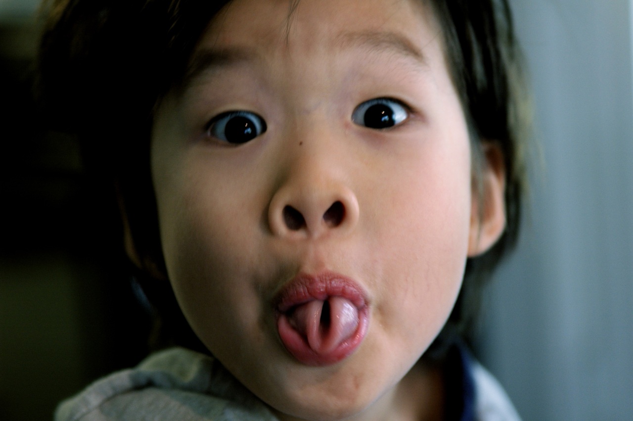 This is an image of a young boy’s face on a blurred background. His rolled tongue is protruding from his mouth.