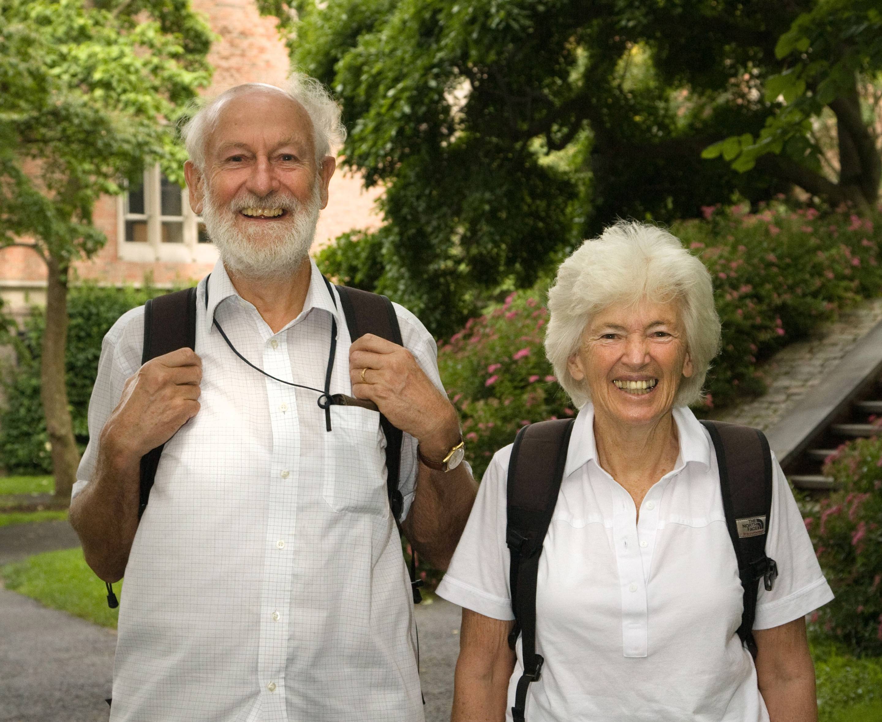 This is an image of two people, a man and a woman, standing side by side and looking at the camera and smiling. They are both wearing white buttoned shirts and a backpack with black straps. Both have grey hair, and the man on the left has a grey beard. The background is trees with a red-brick building in the distance. A concrete staircase runs up behind the woman on the right of the image.