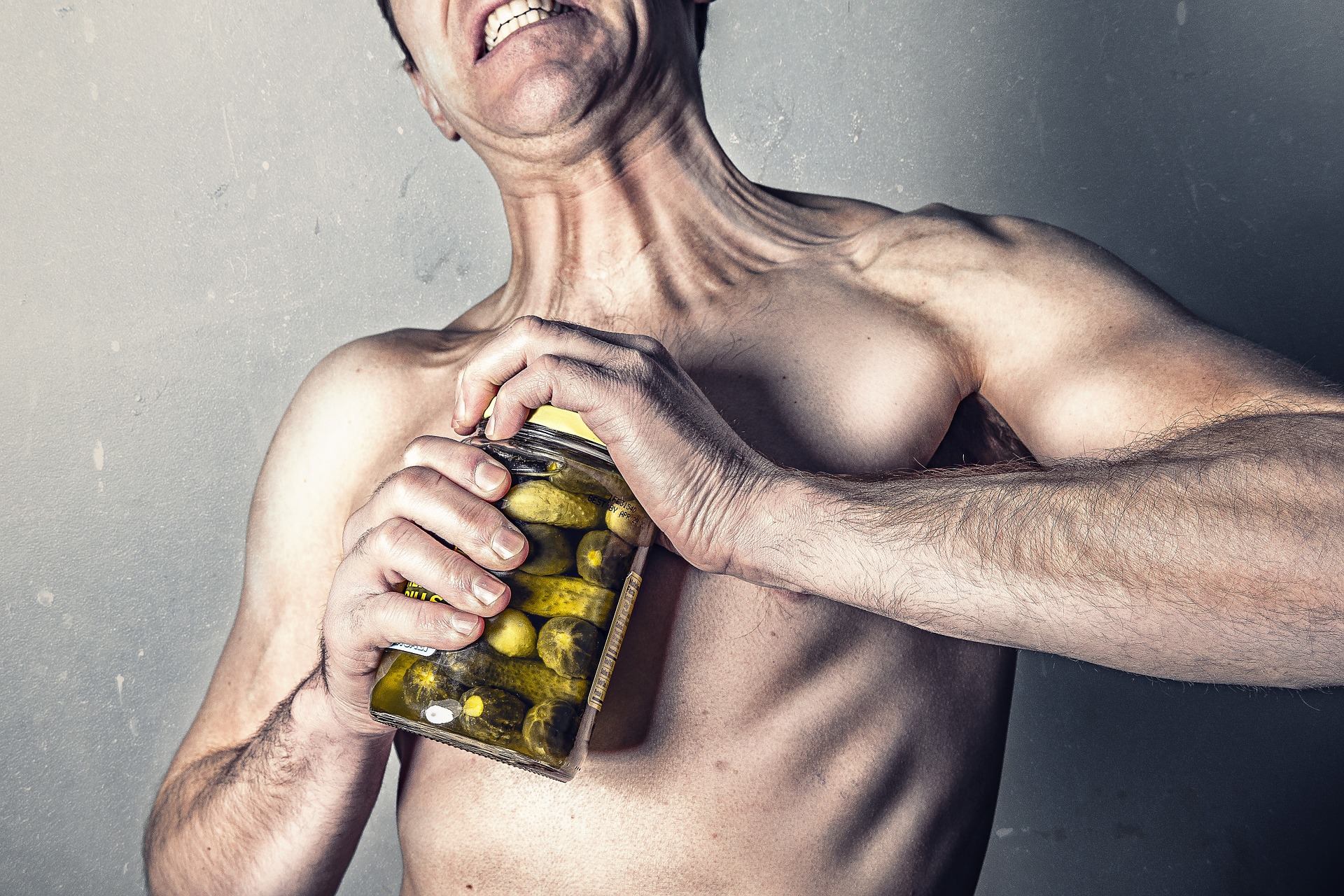 This is an image of a man struggling to open a glass jar of green pickles. The image shows him from the nose down to the waist. He is not wearing a shirt, and he exerting lots of effort to open the jar based on his flexed muscles and bared teeth. The image has a grey background.